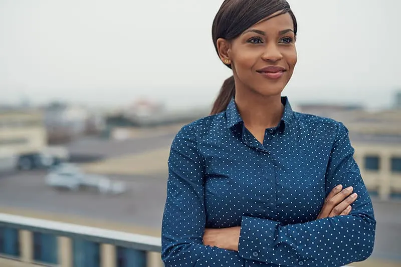 Confident friendly black business woman standing with folded arms on the rooftop of an urban commercial building smiling as she looks to the side of the camera 