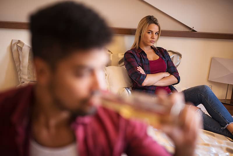 Angry woman sitting on bed behind her drunk boyfriend.