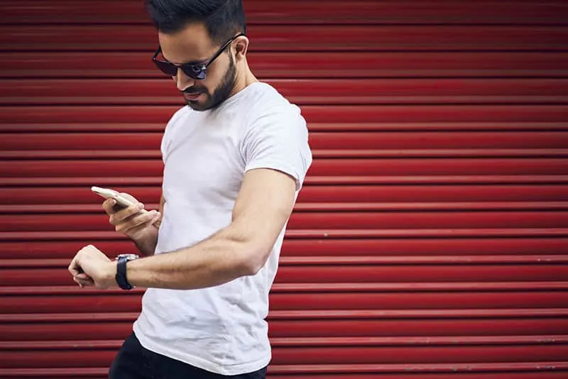 Handsome young bearded man in eyewear and casual outfit receiving text mail with invitation for meeting in cafe strolling outdoors hurrying to get there in time passing copy space for advertising