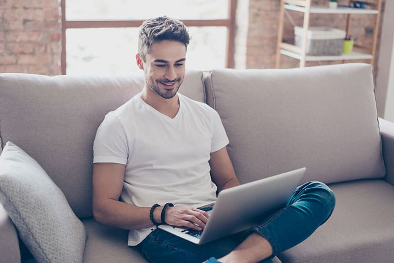 Giovane attraente ragazzo sorridente sta navigando al suo computer portatile, seduto a casa sul divano accogliente beige a casa, indossando abiti casual