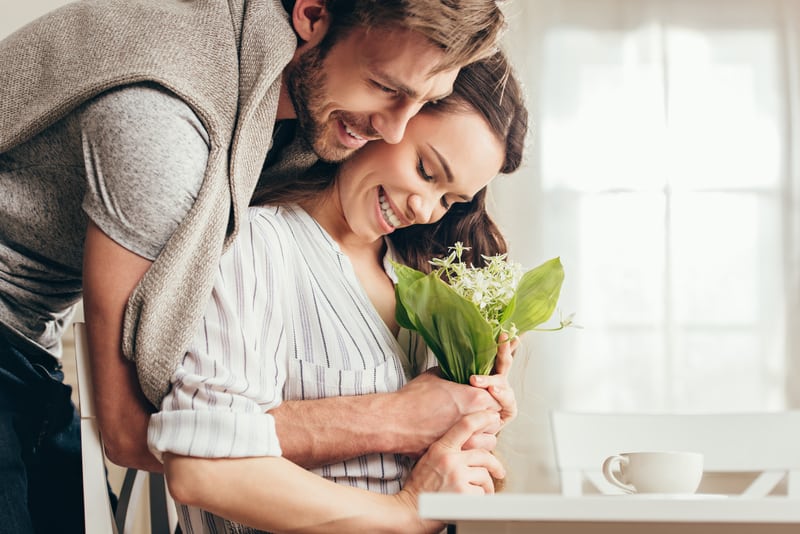 giovane coppia sorridente che si abbraccia e tiene in mano un mazzo di fiori a casa propria