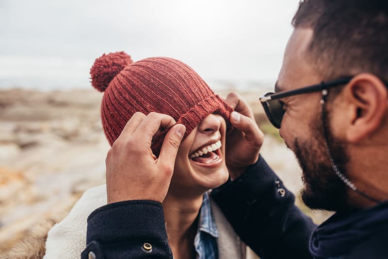 Loving couple having fun outdoors. Man covering eyes of woman with cap