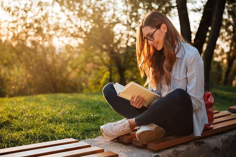 Vista lateral de mujer morena complacida con gafas sentada en un banco y leyendo un libro en un parque.