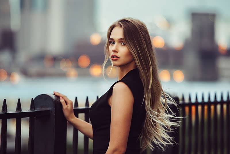Splendida giovane donna modello con perfetti capelli biondi che guarda la fotocamera in posa in città indossando un abito da sera nero