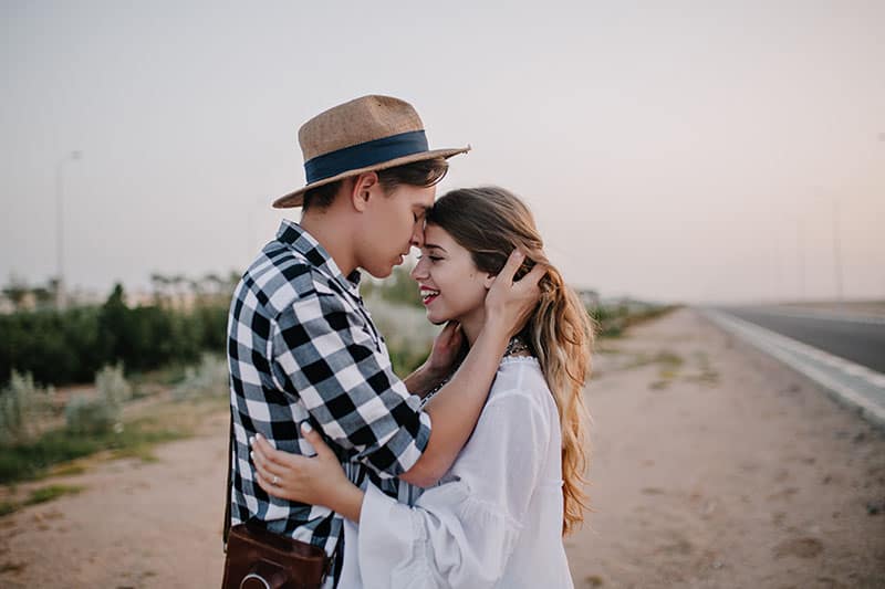 Excited cheerful girl in white tunic soft embracing boyfriend in stylish shirt, while he touching her long hair