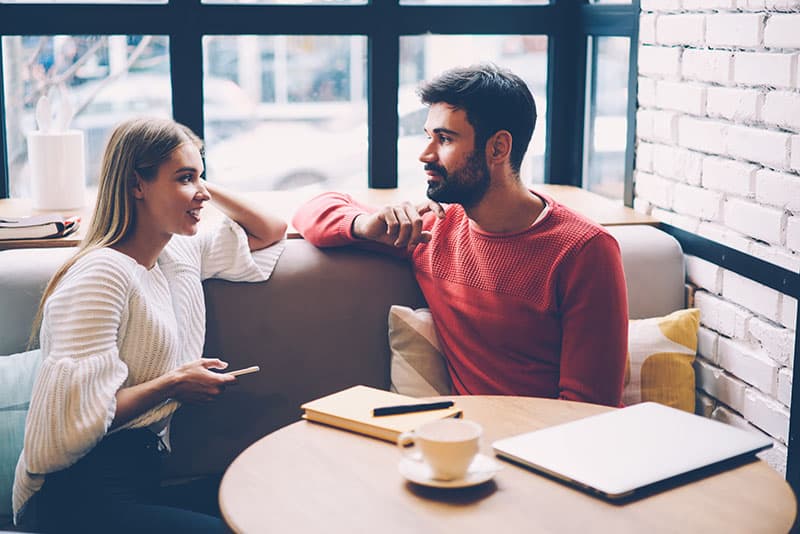 Young woman and man talking during first date in cozy coffee shop enjoying free time together,hipster girl checking notification on mobile while spending time with boyfriend having lovely conversation