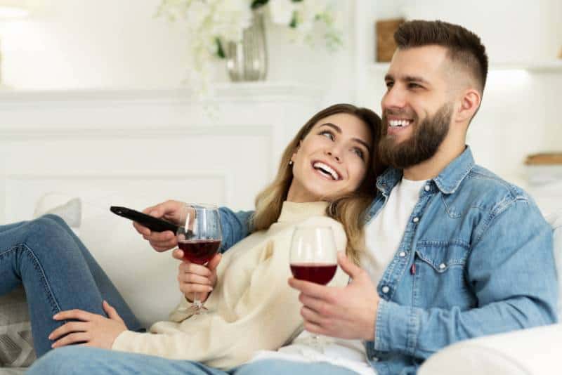 smiling couple watching TV while holding glass of wine in living room