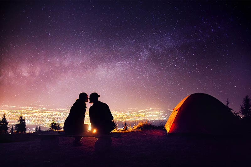 Casal feliz em silhueta a beijar-se perto de uma fogueira e de uma tenda cor de laranja