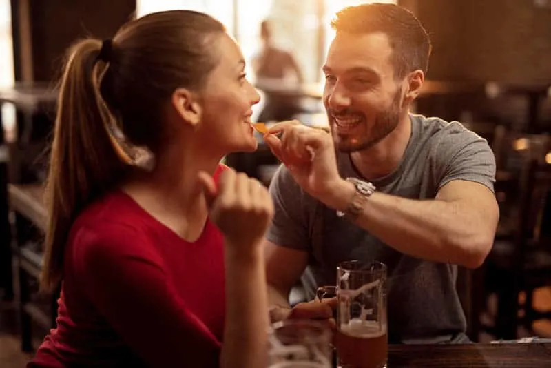 Happy man feeding his girlfriend with nacho chips while drinking beer together in a pub