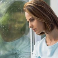 sad woman standing by the window while rain is falling down