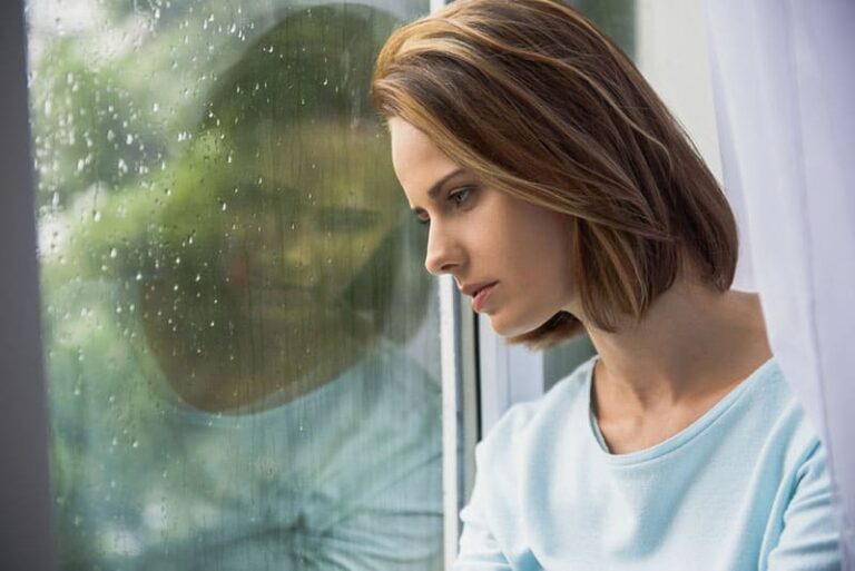 sad woman standing by the window while rain is falling down