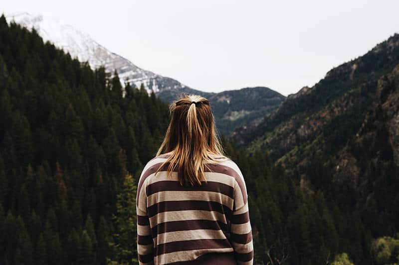 back view of blonde girl wearing long sleeve shirt and standing in front of mountains
