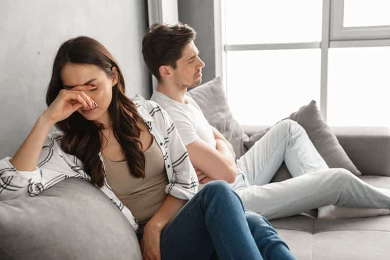 Foto di coppia delusa seduta insieme sul divano di casa con sguardo sconvolto ed espressione di litigio isolata su sfondo bianco