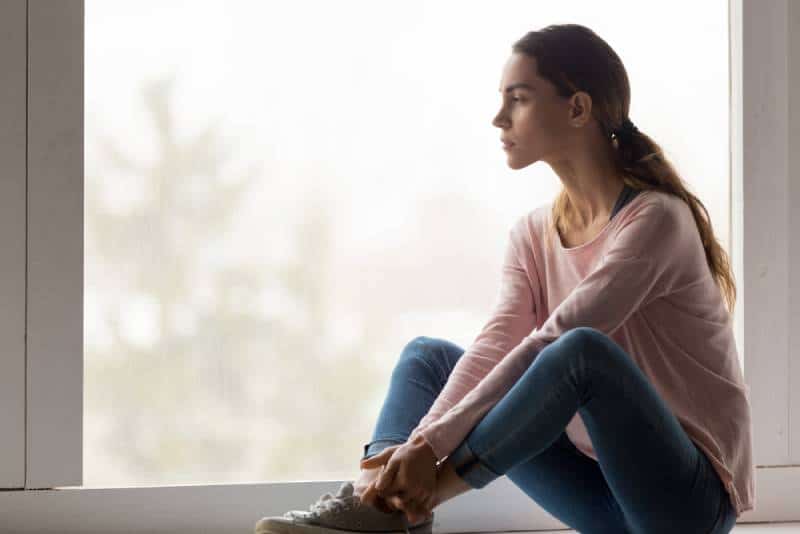 Sad thoughtful girl sit alone on sill looking through window