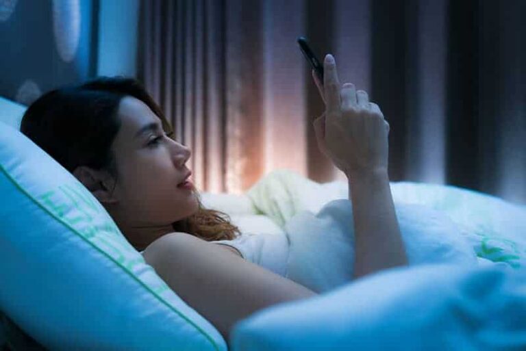 vista lateral de uma mulher deitada na cama e a escrever no telemóvel durante a noite