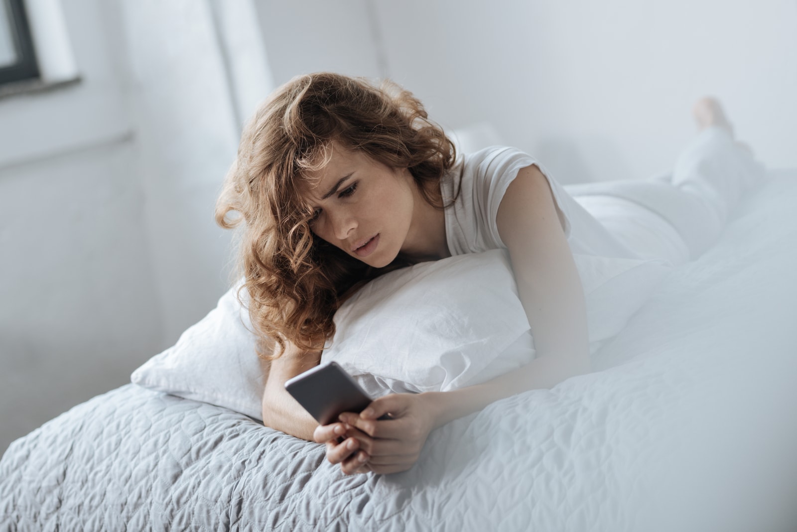 una bionda triste sdraiata sul letto che usa uno smartphone