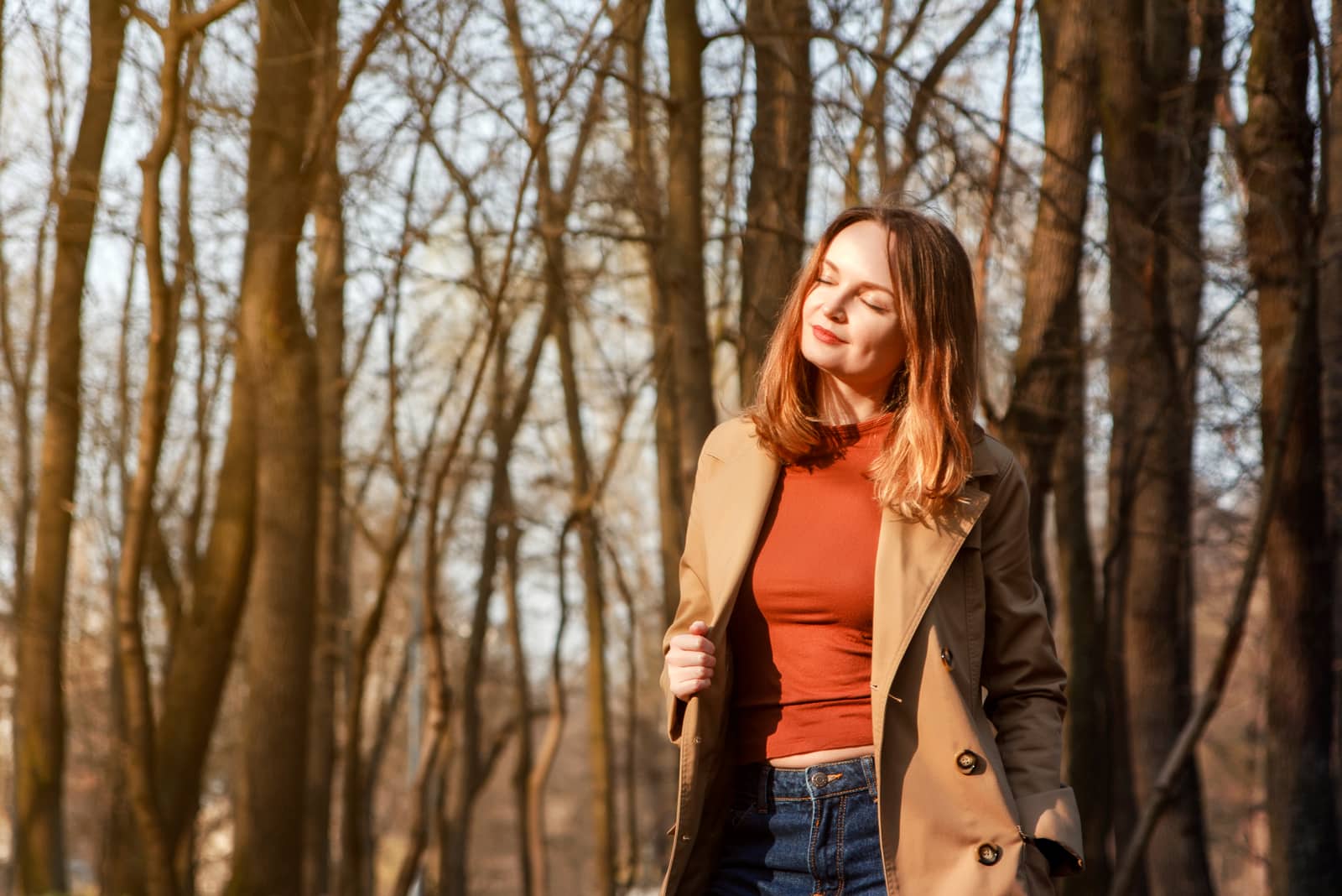 a smiling brunette in the woods stands in a brown coat