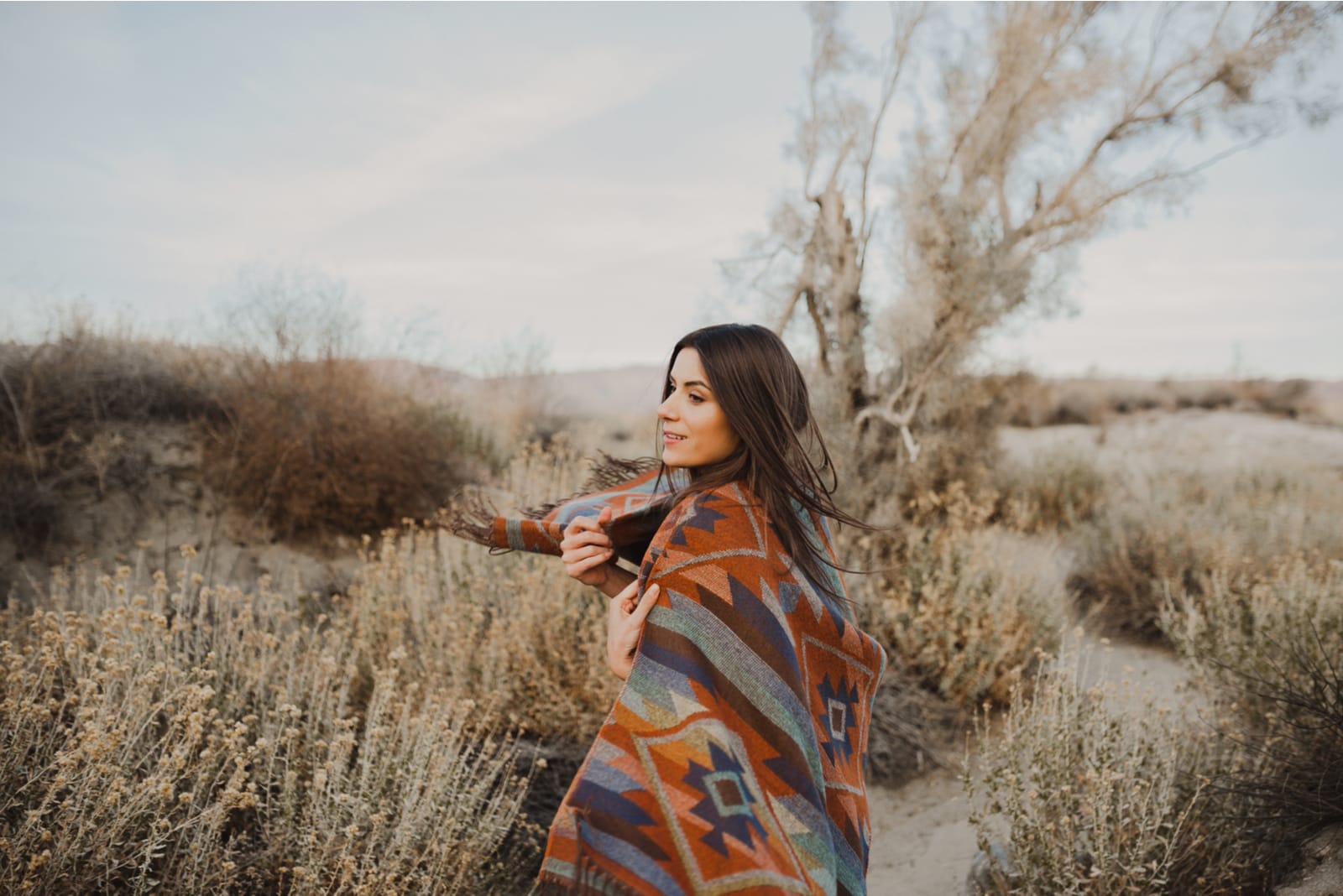 a smiling woman standing in nature wrapped in a brown cloak