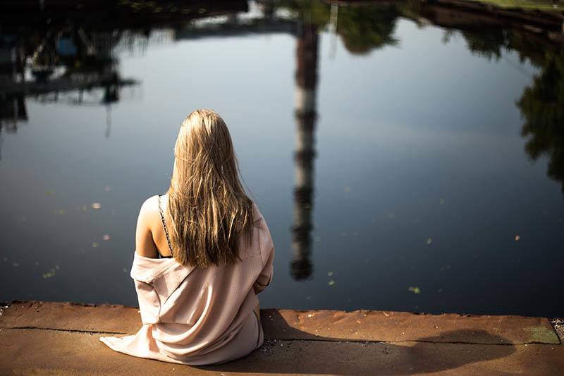vista posteriore di una donna bionda seduta su un pontile di legno davanti a un lago