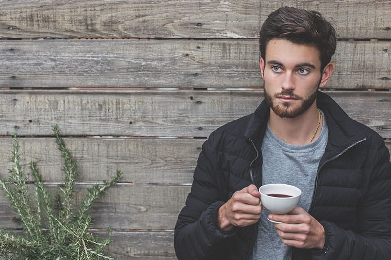 man holding a cup of coffee while leaning on wooden wall