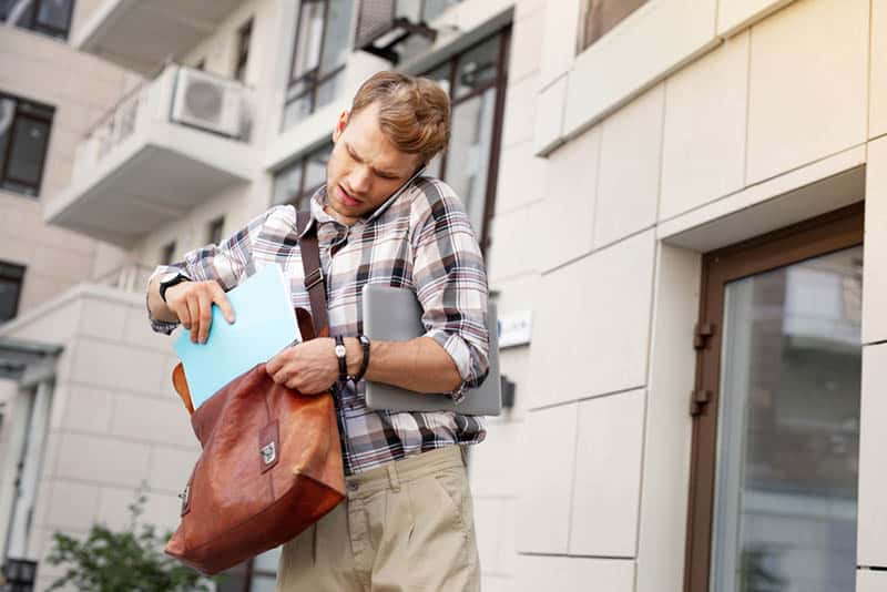 man putting book in bag in front of building