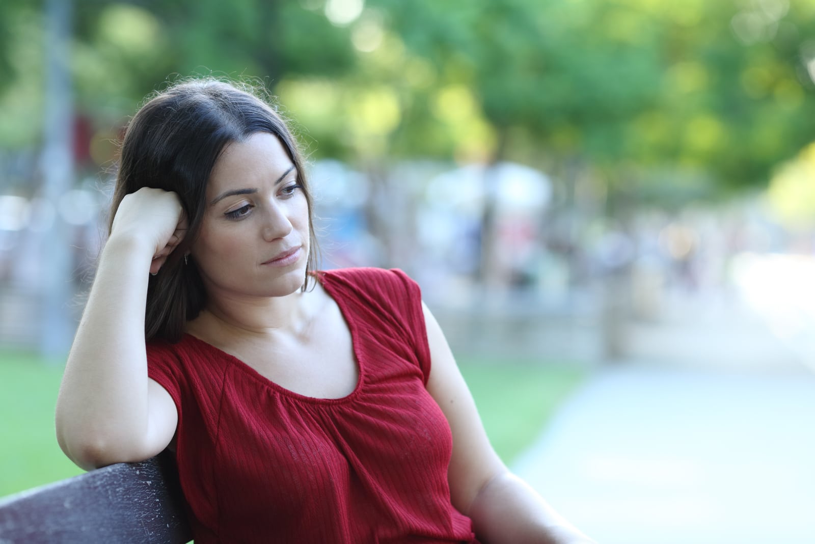 mujer pensativa con camiseta roja sentada en un banco del parque