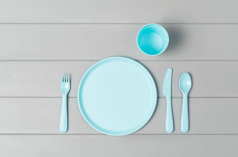 Set of empty, pastel plastic tableware bowls, spoons, knifes and forks isolated on gray wooden background. Flat lay, top view with minimal style.
