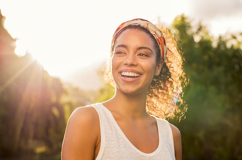 Ritratto di bella donna afroamericana che sorride e guarda lontano al parco durante il tramonto. Ritratto all'aperto di una ragazza nera sorridente. Ragazza felice e allegra che ride al parco con fascia per capelli colorata. 