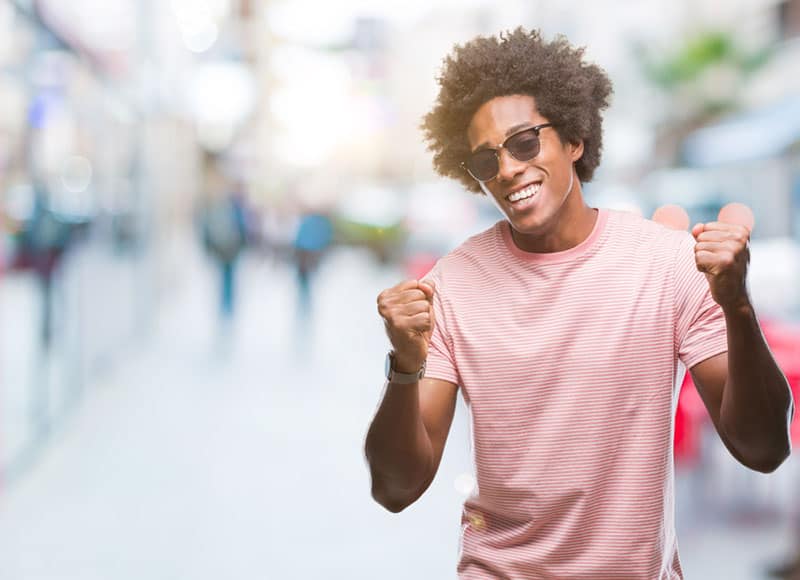 Homem afro-americano com óculos de sol sobre fundo isolado, muito feliz e entusiasmado, a fazer um gesto de vencedor com os braços levantados, a sorrir e a gritar pelo sucesso. Conceito de celebração