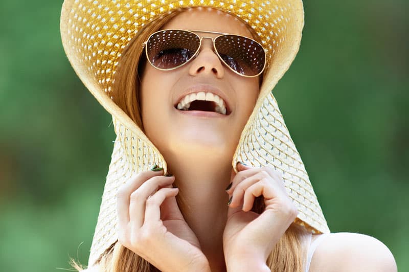 Retrato de uma jovem sorridente com um chapéu de praia largo, tendo como fundo um parque verde de verão.