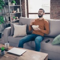 Portrait of his he nice-looking attractive bearded cheerful cheery positive guy spending weekend sitting on divan watching tv at industrial loft interior style living-room indoors