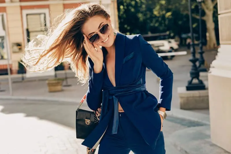 attractive smiling woman with long hair walking on sunny weather in autumn city wearing sexy blue suit, street fashion style