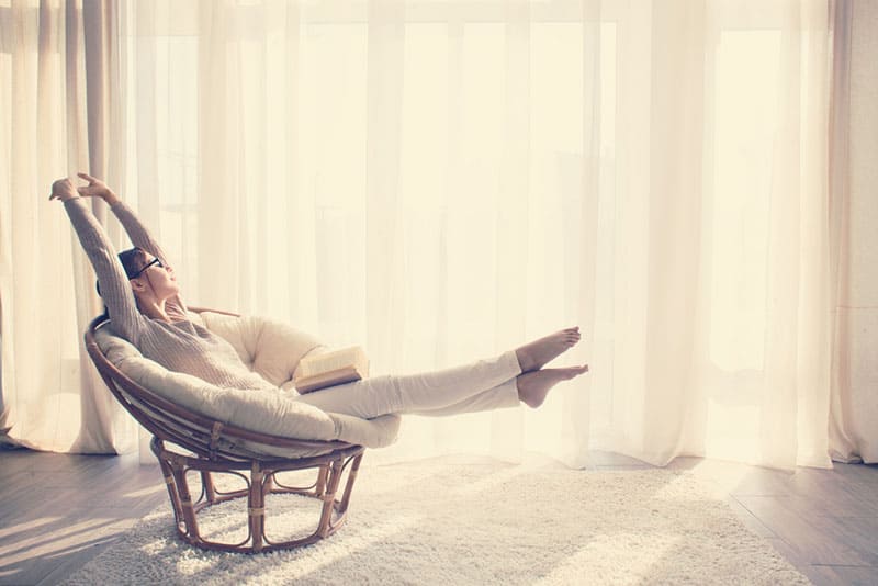 Giovane donna a casa seduta su una sedia moderna di fronte alla finestra che si rilassa nel suo salotto leggendo un libro, instagram tonificazione