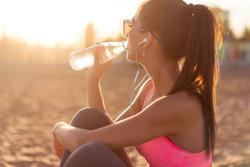 Bella donna atleta fitness bere acqua dopo il lavoro di esercizio sul tramonto sera estate in spiaggia ritratto all'aperto