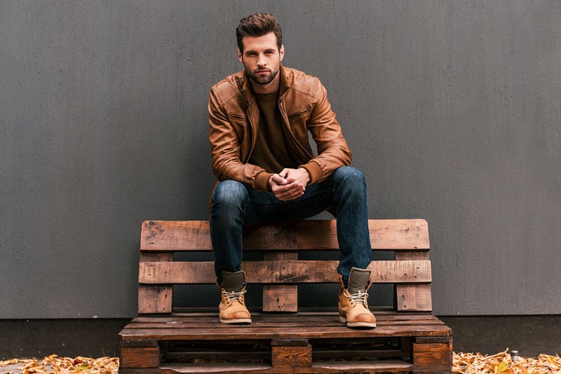 Seguro de sí mismo y guapo. Joven guapo sentado en el palé de madera y mirando a cámara con pared gris de fondo y hojas caídas naranjas en el suelo.