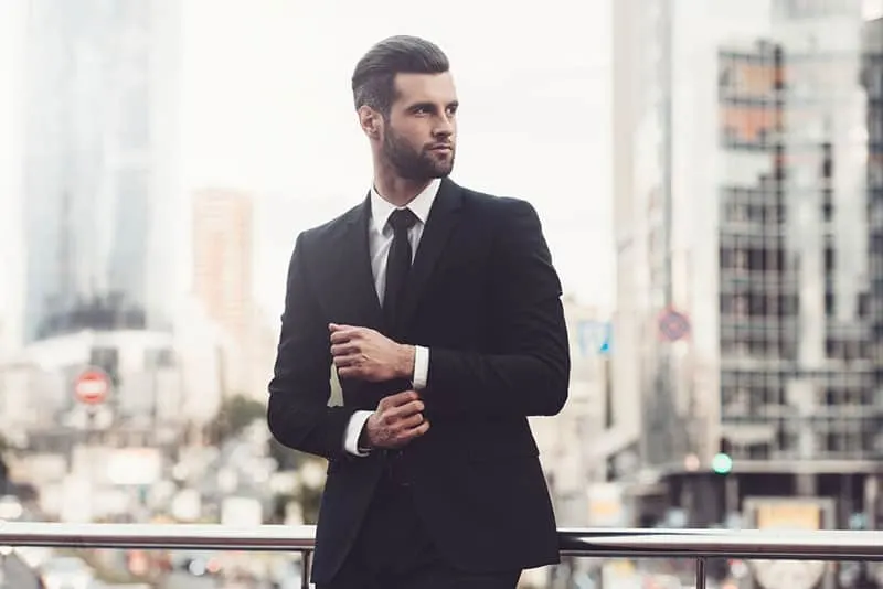Modern businessman. Confident young man in full suit adjusting his sleeve and looking away while standing outdoors with cityscape in the background