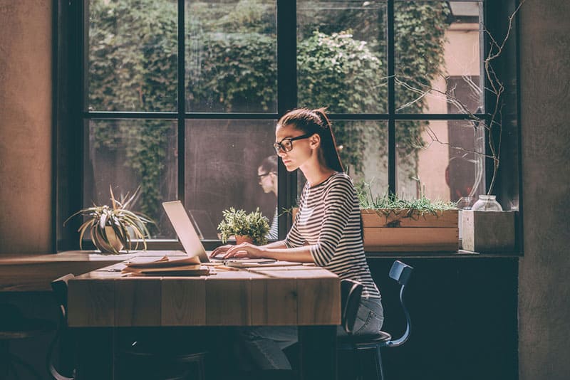 Concentrata al lavoro. Giovane donna sicura di sé in abbigliamento casual elegante che lavora al computer portatile mentre è seduta vicino alla finestra di un ufficio creativo o di un caffè.