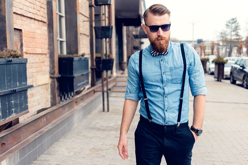 Portrait of a brutal bearded man walking in the street. Young stylish hipster posing, sunglasses, mustache, the bow tie, suspenders. Businessman looking looking at the camera outdoors.