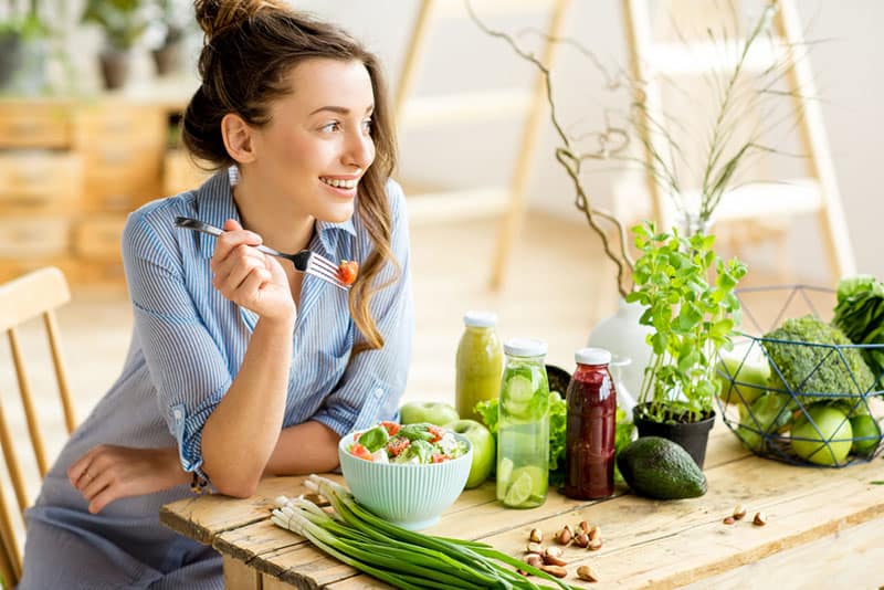 Donna giovane e felice che mangia un'insalata sana seduta sul tavolo con ingredienti verdi e freschi all'aperto 