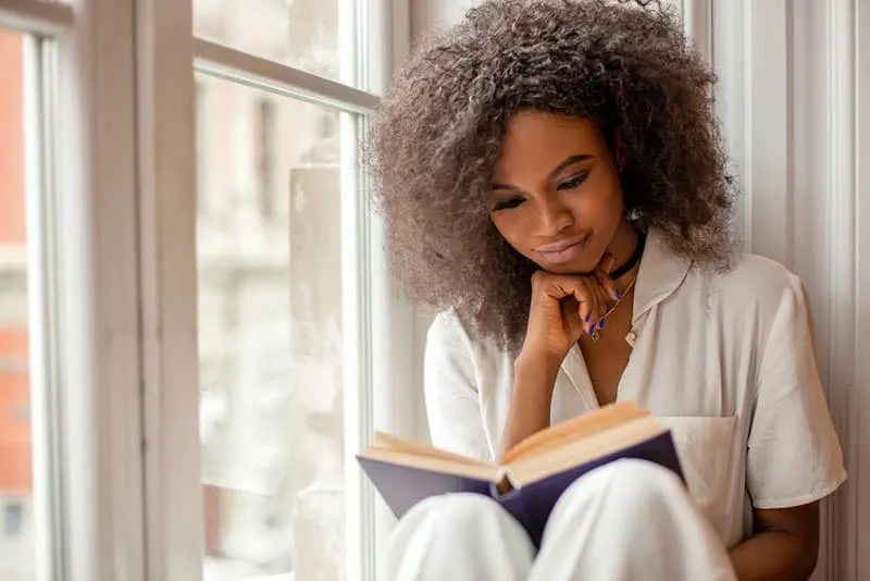 Pretty afro-american girl with reading a book sitting on the windowsill.