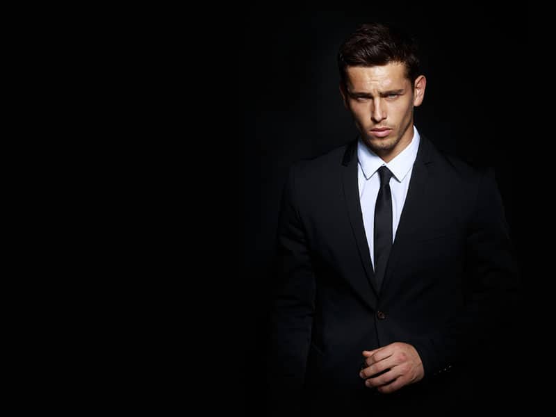 Handsome young brunette model, wearing in black and white suit, looking with attitude, posing at studio, isolated on black background. Business man portrait. Copy space. Horizontal view.