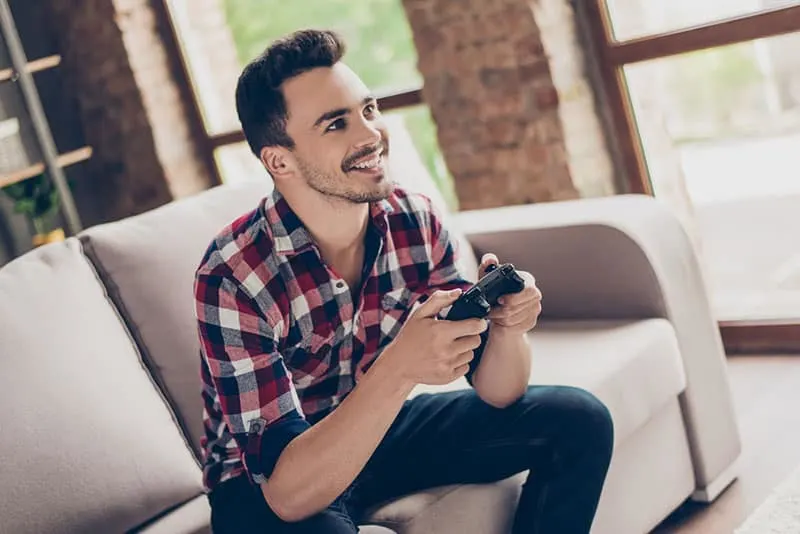Portrait of attractive brunet hipster with bristle, holding joy stick and playing videogames on tv at vacation, sits at home on the cozy couch, successful, he is winning at crazy car race