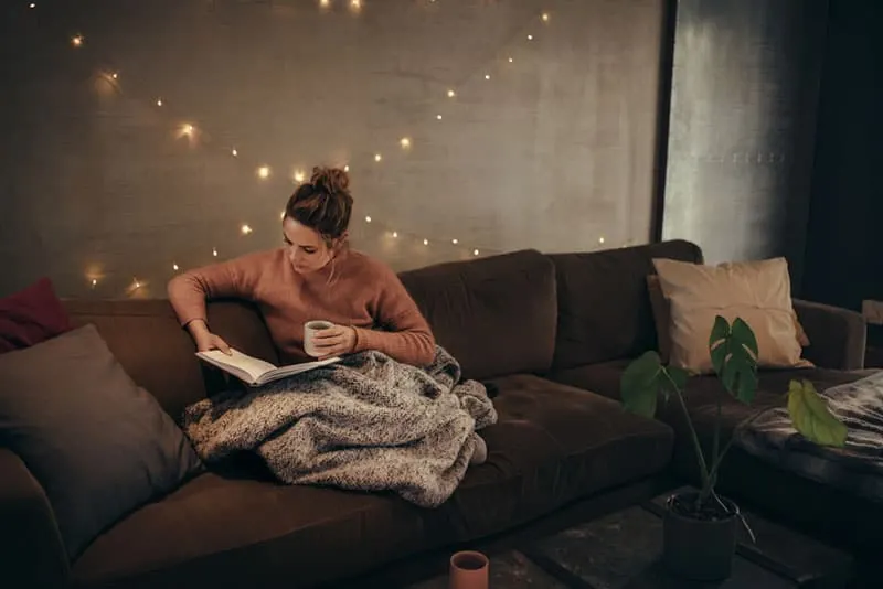 Young woman reading book and drinking coffee on sofa in hygge house. Caucasian female relaxing in cozy living room and reading a book.