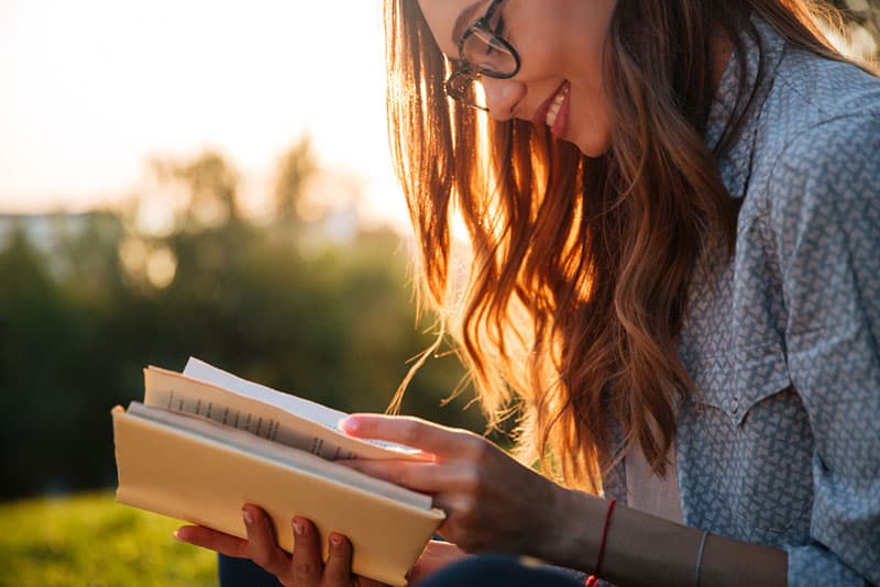 Imagen recortada de una mujer morena sonriente con gafas leyendo un libro en un parque.