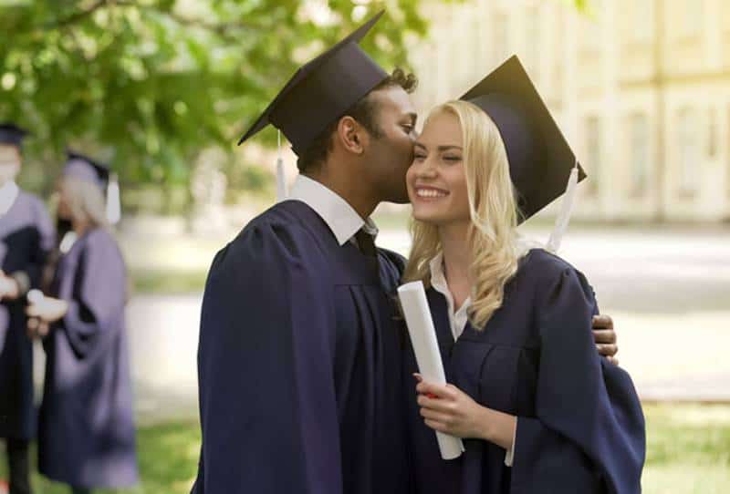 Graduados en trajes académicos sonriendo, chico feliz besando a su novia en la mejilla