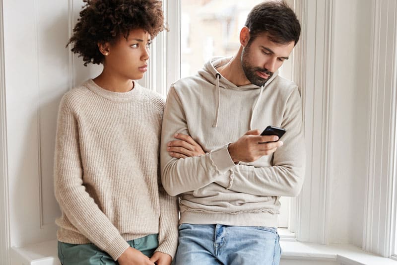 Sad African woman in oversized sweater feels displeased as being neglegted by boyfriend, needs lively communication, her husband addicted to modern technologies. Family relationships concept