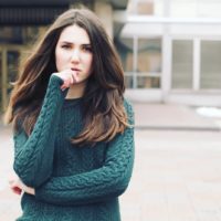 a sad brunette in a green sweater stands outside