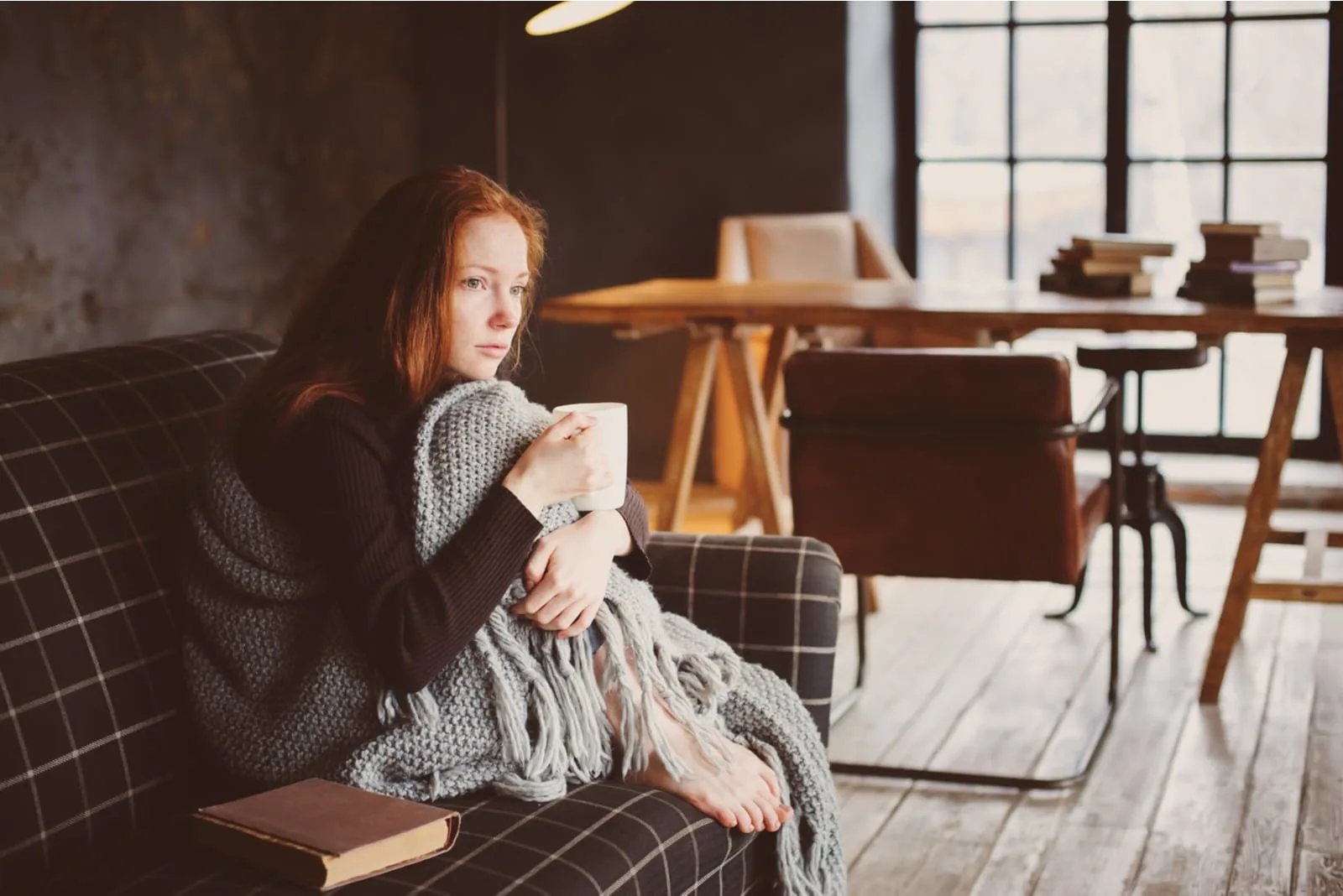 a sad red-haired girl sits on the couch and drinks tea