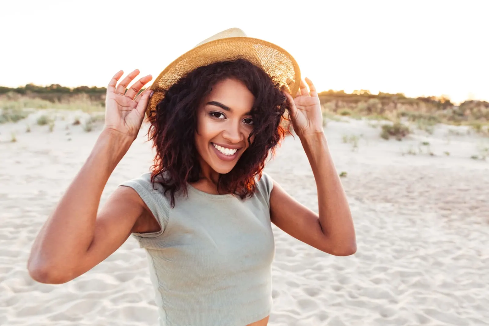 a smiling black woman with a hat stands on the beach