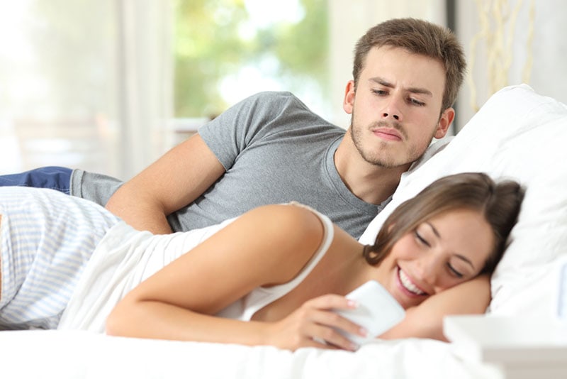 jealous man looking at woman typing in the bed